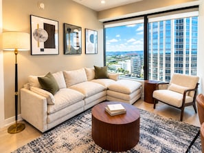 A modern living room with a sectional sofa, a round coffee table, and a chair. There are framed artworks on the wall and a large window with a city view. - The Birmingham Heights | 2BR Residence (Birmingham)