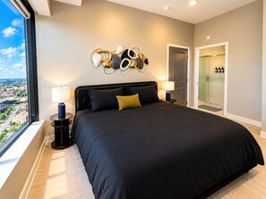A modern bedroom with a large bed, two bedside lamps, a city view, a door with a glass panel, and a wall art piece. - The Birmingham Heights | 2BR Residence (Birmingham)