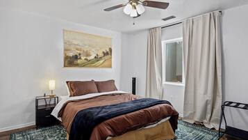 Un dormitorio con cama, un cuadro, una mesita de noche, una lámpara, una ventana con cortinas y un ventilador de techo.