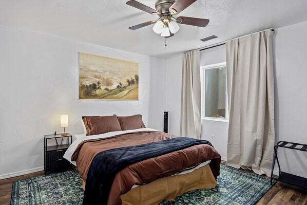 A bedroom with a bed, a painting, a nightstand, a lamp, a window with curtains, and a ceiling fan.