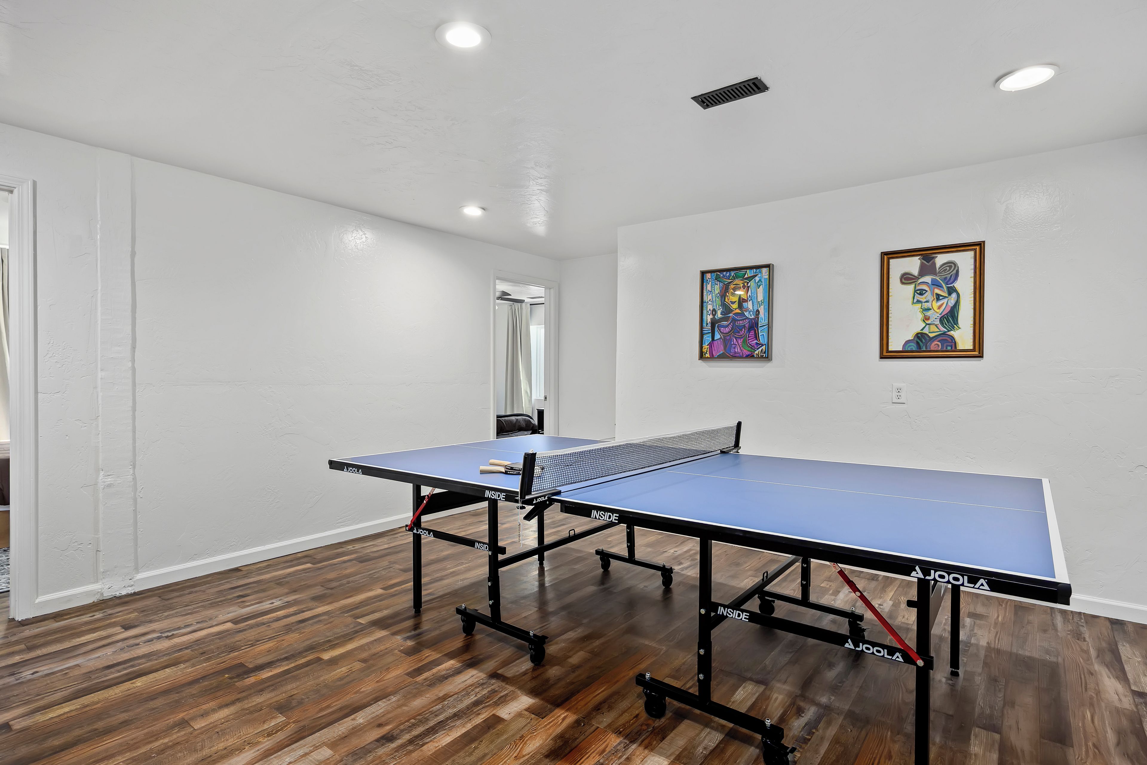 A room with a ping pong table, wooden flooring, white walls, and two paintings.