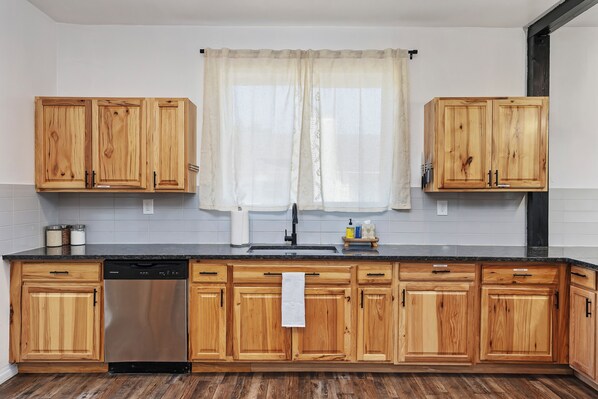 A kitchen with wooden cabinets, a stainless steel dishwasher, and a window with a sheer curtain.