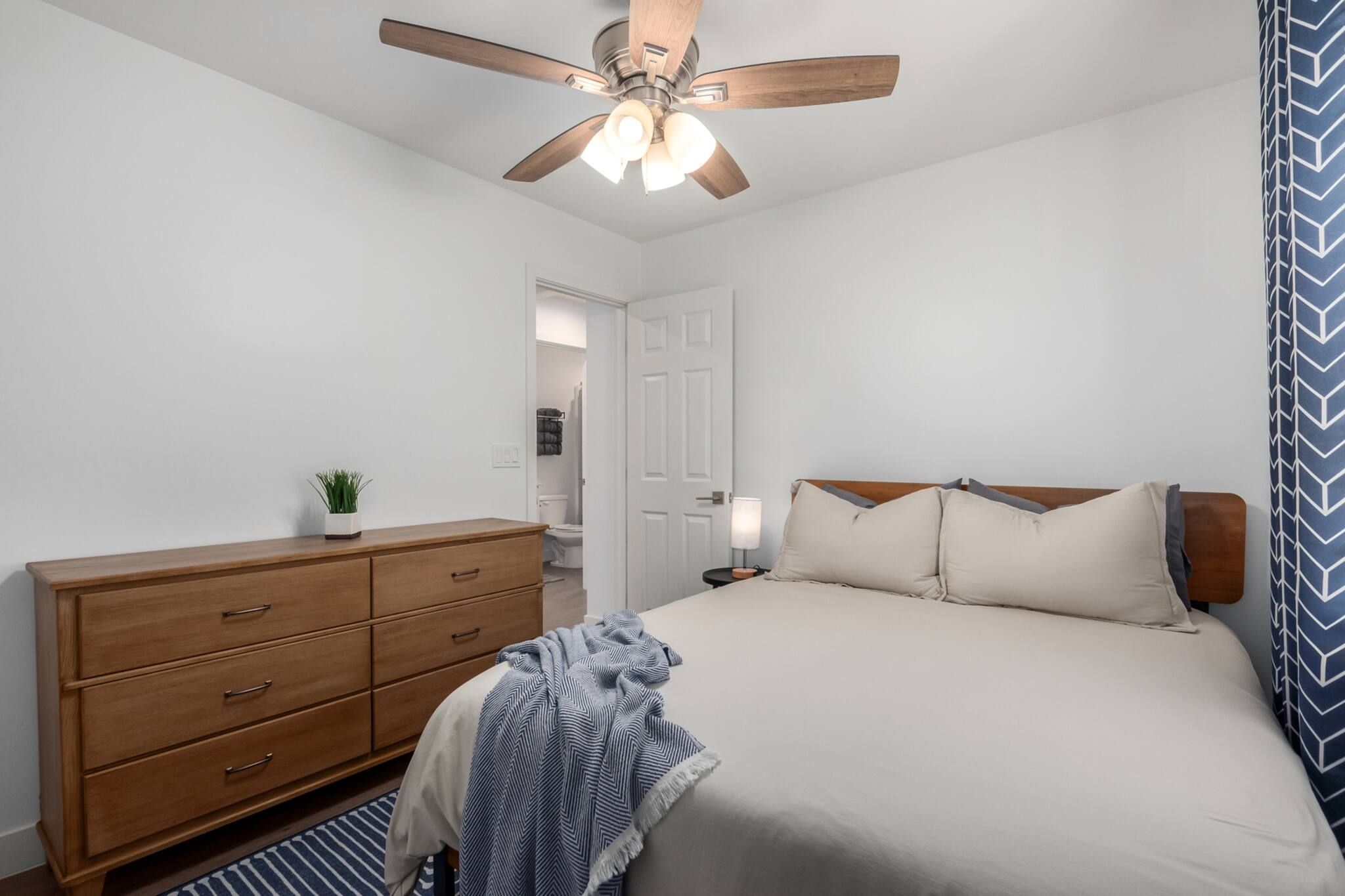 A bedroom with a bed, wooden dresser, ceiling fan, and a door leading to another room.