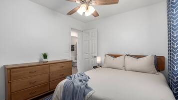 A bedroom with a bed, wooden dresser, ceiling fan, and a door leading to another room.
