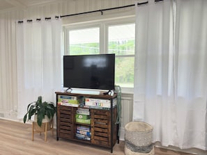 Une pièce avec un téléviseur posé sur un meuble en bois, une plante en pot et une fenêtre avec des rideaux.