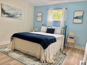 A bedroom with a bed, a nightstand, a window with curtains, and artwork on the walls.