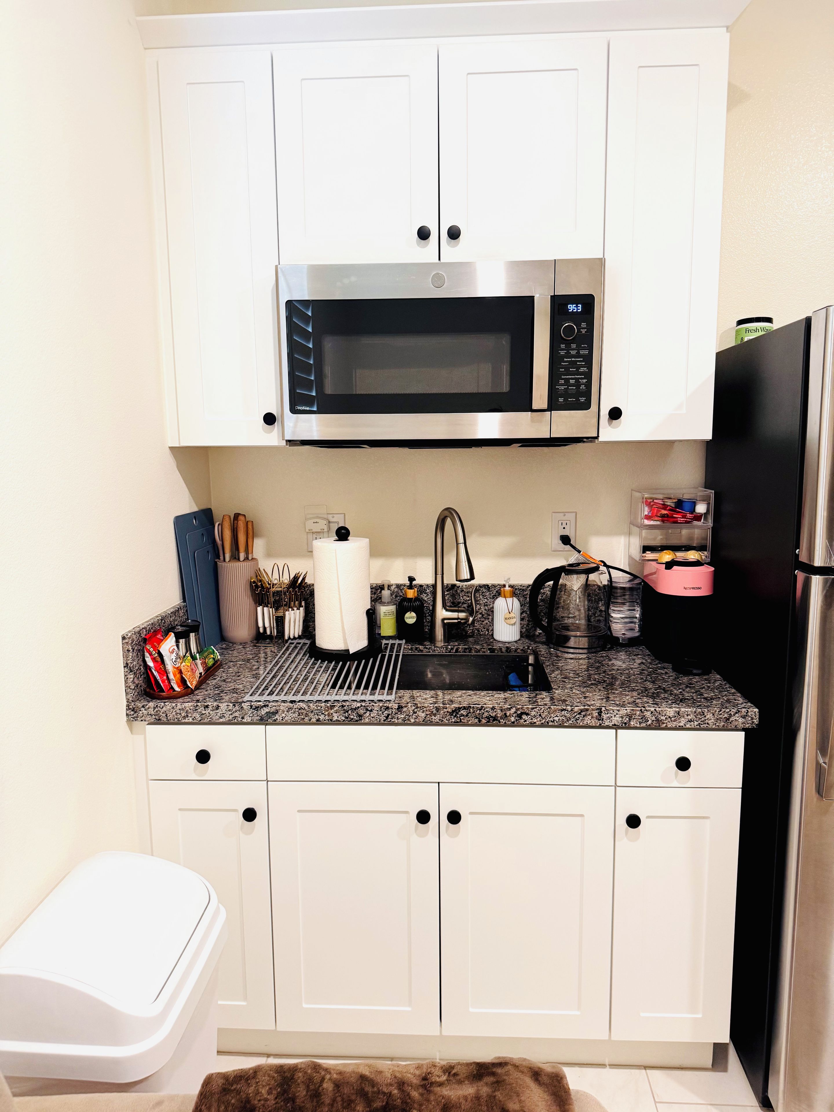 A compact kitchen with white cabinets, a stainless steel microwave, a black refrigerator, and a granite countertop.