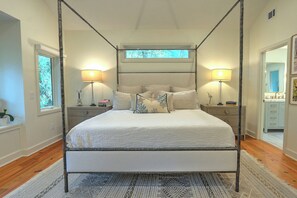 A four-poster bed with a canopy, two bedside tables with lamps, a window with a view, and a bathroom with a mirror and sink. - Renovated Sea Pines Retreat with Private Pool (Hilton Head Island)