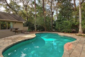A swimming pool with a curved edge, surrounded by a brick deck and a wooden deck with steps leading to a house. - Renovated Sea Pines Retreat with Private Pool (Hilton Head Island)