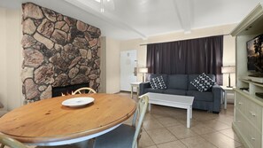 Ein Wohnzimmer mit Kamin aus Stein, einer Couch mit gemusterten Kissen, einem Fernseher und einem Essbereich mit Tisch und Stühlen.