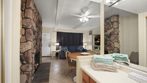 A living room with a stone wall, a fireplace, a ceiling fan, a sofa, a dining table, and a television. - Palm Springs Fireside Retreat | King Bed + Pool (Palm Springs)