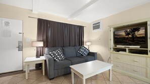 A living room with a grey sofa, two patterned pillows, a white coffee table, a TV on a cabinet, and a door with a key card reminder. - Palm Springs Fireside Retreat | King Bed + Pool (Palm Springs)