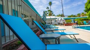 A row of blue lounge chairs by a poolside, with a building featuring vertical windows in the background. - Desert Days & Poolside Ways | 1BR Suite (Palm Springs)