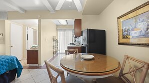 A dining area with a round glass table, chairs, a refrigerator, and a painting on the wall. - Desert Dreamin’ Studio | King Bed + Pool (Palm Springs)