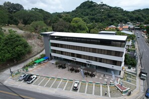 A multi-story building with a flat roof, a parking area with marked spaces, and a street view with vehicles and greenery in the background. - Hotel Plaza Iguana (Quepos)