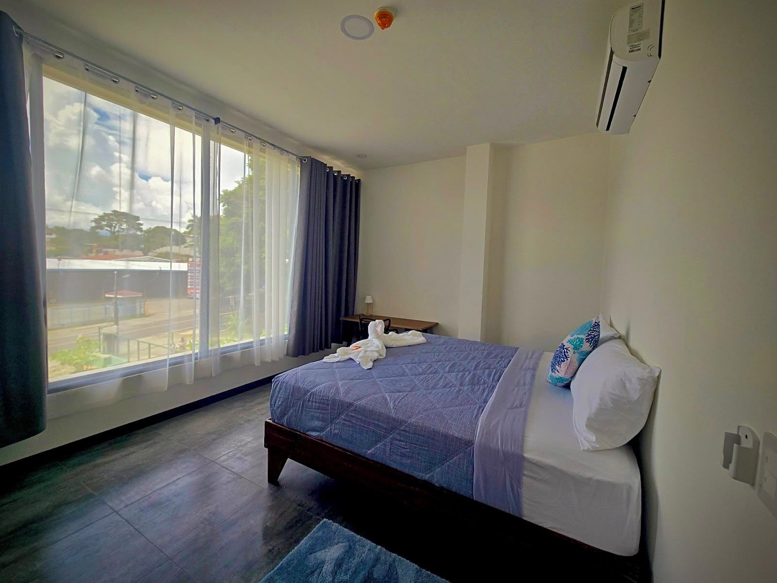 A hotel room with a bed, a window with curtains, a small table, and a wall-mounted air conditioning unit.