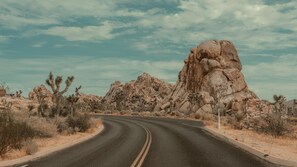 Estrada no deserto serpenteia por uma paisagem rochosa, com vegetação rala e céu claro.
