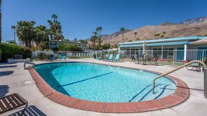A swimming pool with a brick edge, lounge chairs, and a building with a blue roof in the background. - Desert Charm & Fireside Comfort (Palm Springs)