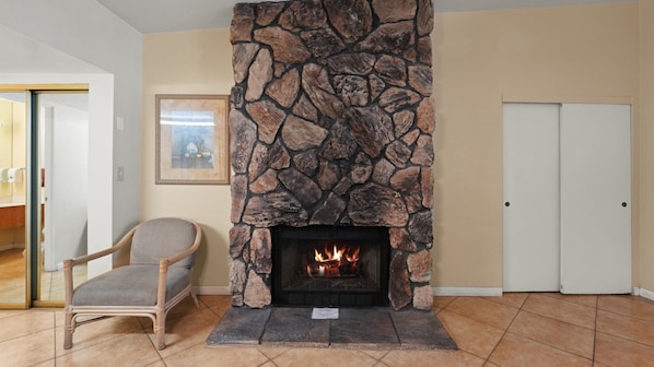 A stone fireplace with a fire, a wicker chair, and a framed picture on the wall. - Desert Charm & Fireside Comfort (Palm Springs)