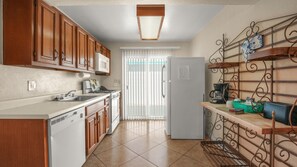 A kitchen with wooden cabinets, a white refrigerator, a coffee maker, and a metal rack with dishes. - Fireside Palm Springs Escape | 1BR + 2 Baths (Palm Springs)