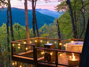 A wooden deck with a hot tub and a grill, surrounded by trees and overlooking a mountainous landscape.