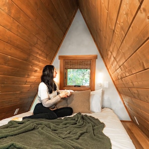 A person sitting on a bed in a triangular wooden room with a window and a wall-mounted light. - A-frame • Hot Tub • Sauna • Outdoor Shower (Penrose)