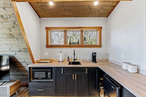 A modern kitchen with a wooden ceiling, a built-in microwave, a sink, and a window with a view of trees. - A-frame • Hot Tub • Sauna • Outdoor Shower (Penrose)
