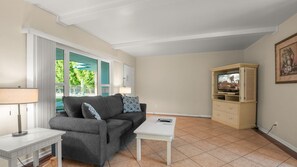 A living room with a grey sofa, a white coffee table, a television on a wooden cabinet, and a window with a view of greenery. - 1 Bedroom for Palm Springs Adventures (Palm Springs)