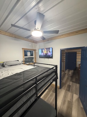 A room with a bunk bed, a television mounted on the wall, a ceiling fan, and a door leading to another room.