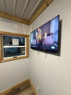 A room with a large flat-screen TV mounted on the wall, a window with a wooden frame, and a wooden ceiling.