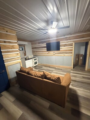 Una habitación con techo de madera, un televisor de pantalla plana montado en la pared, una kitchenette con estufa y horno, y un sofá marrón con cojines.