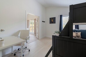 Ein modernes Zimmer mit weißem Schreibtisch, einem Sessel, einem fenster mit Vorhängen, einer Pflanze und einer Couch mit Kissen.