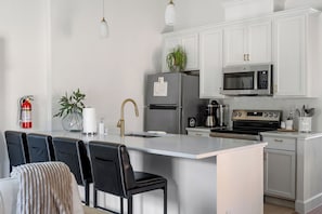 Eine moderne Küche mit weißen Schränken, einem Edelstahl-Kühlschrank, einer Mikrowelle und einer Kaffeemaschine. Es sind Sitzhocker und ein Feuerlöscher zu sehen.