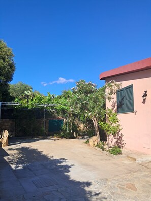 A courtyard with a stone floor, a pink building with a green window, and a pergola covered with green fabric. - Solanas_A cozy and relaxing villa with plenty of outdoor space (Cuili Murvoni)