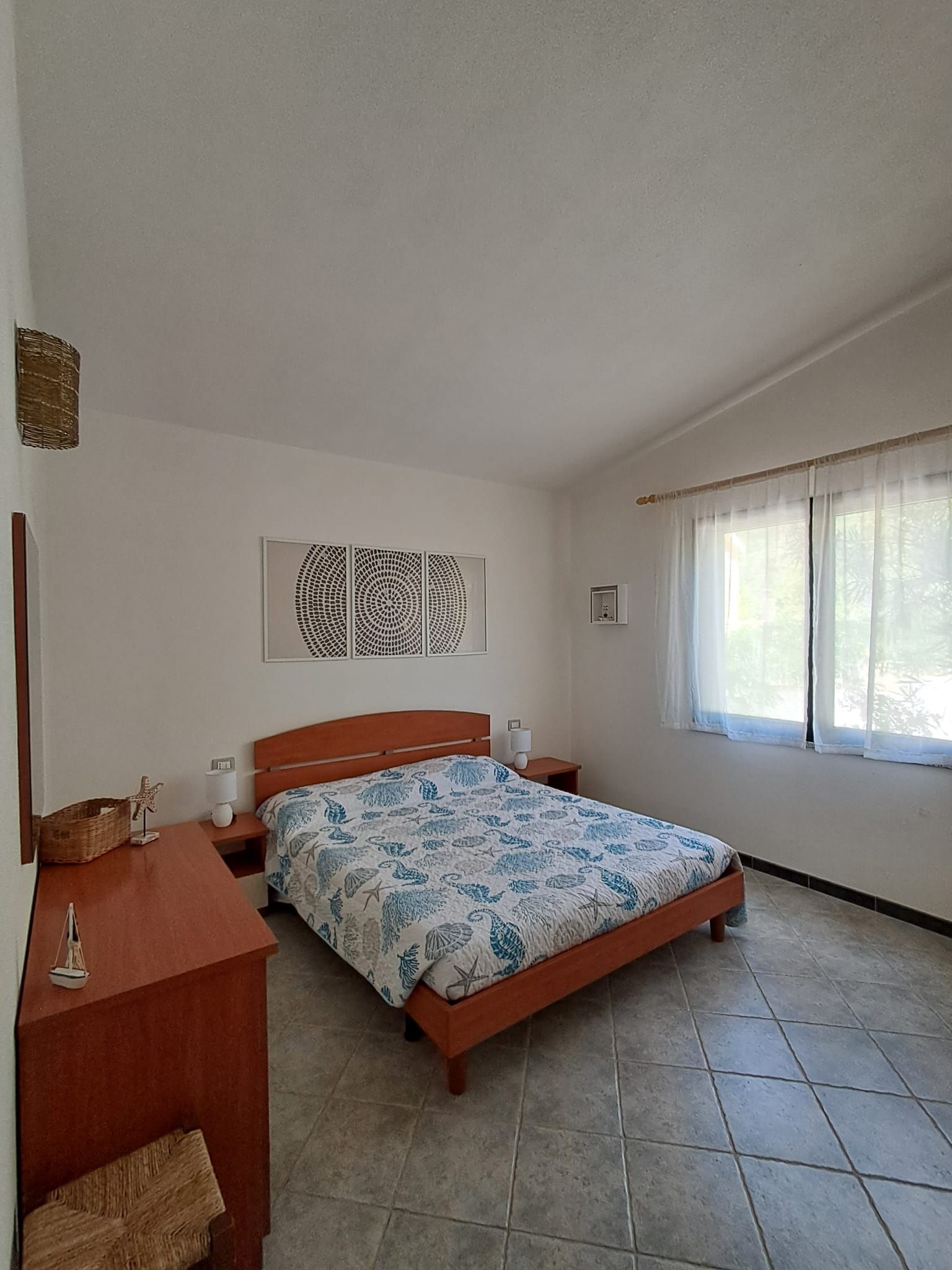 A bedroom with a bed, bedside tables, a wall-mounted light, a window with curtains, and a decorative wall piece.