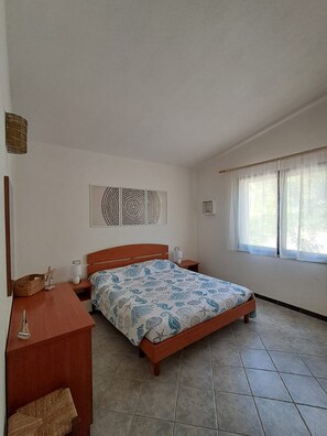 A bedroom with a bed, bedside tables, a wall-mounted light, a window with curtains, and a decorative wall piece. - Solanas_A cozy and relaxing villa with plenty of outdoor space (Cuili Murvoni)