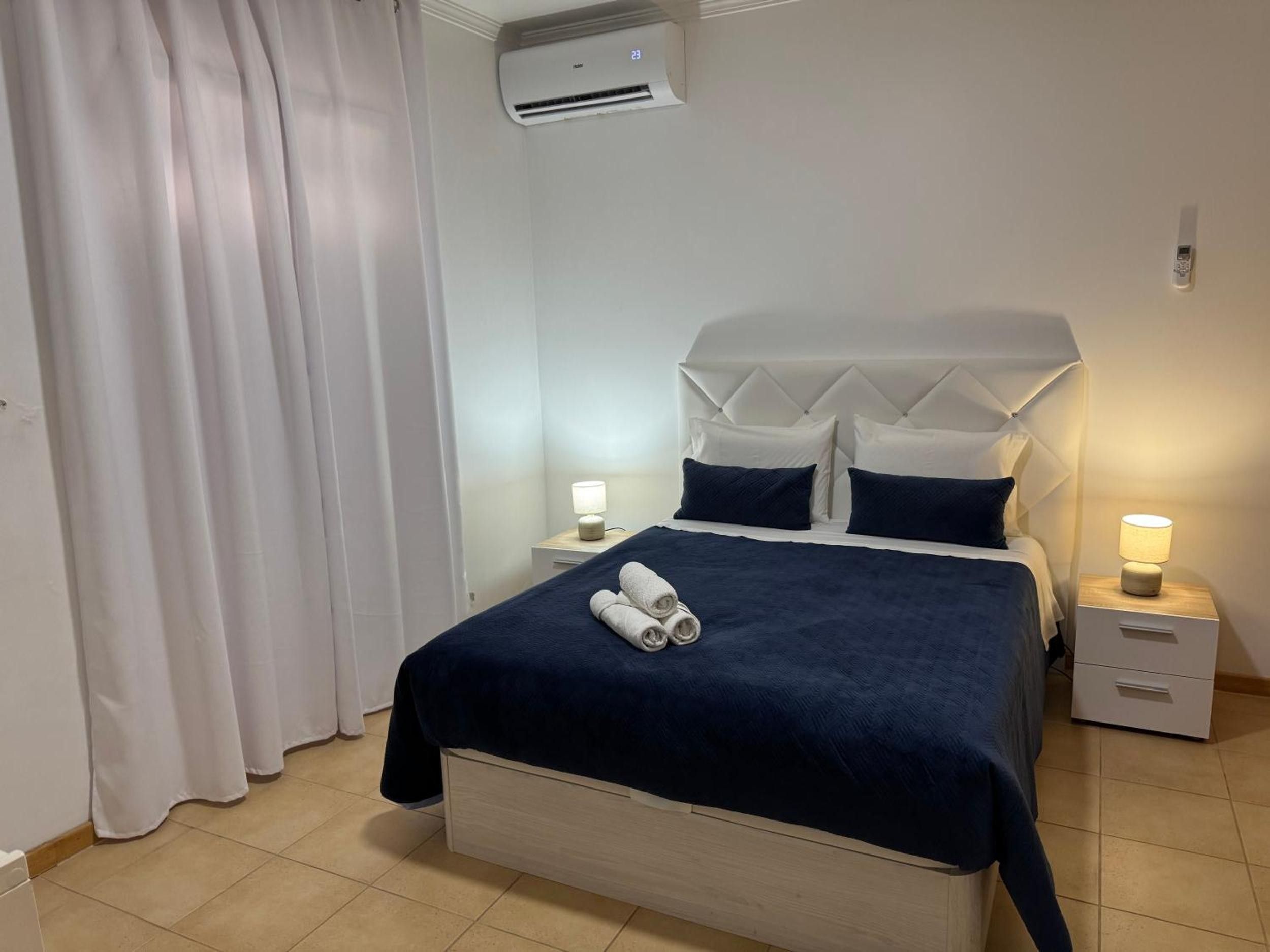 A hotel room with a bed, two bedside tables, a wall-mounted air conditioner, and a curtain.