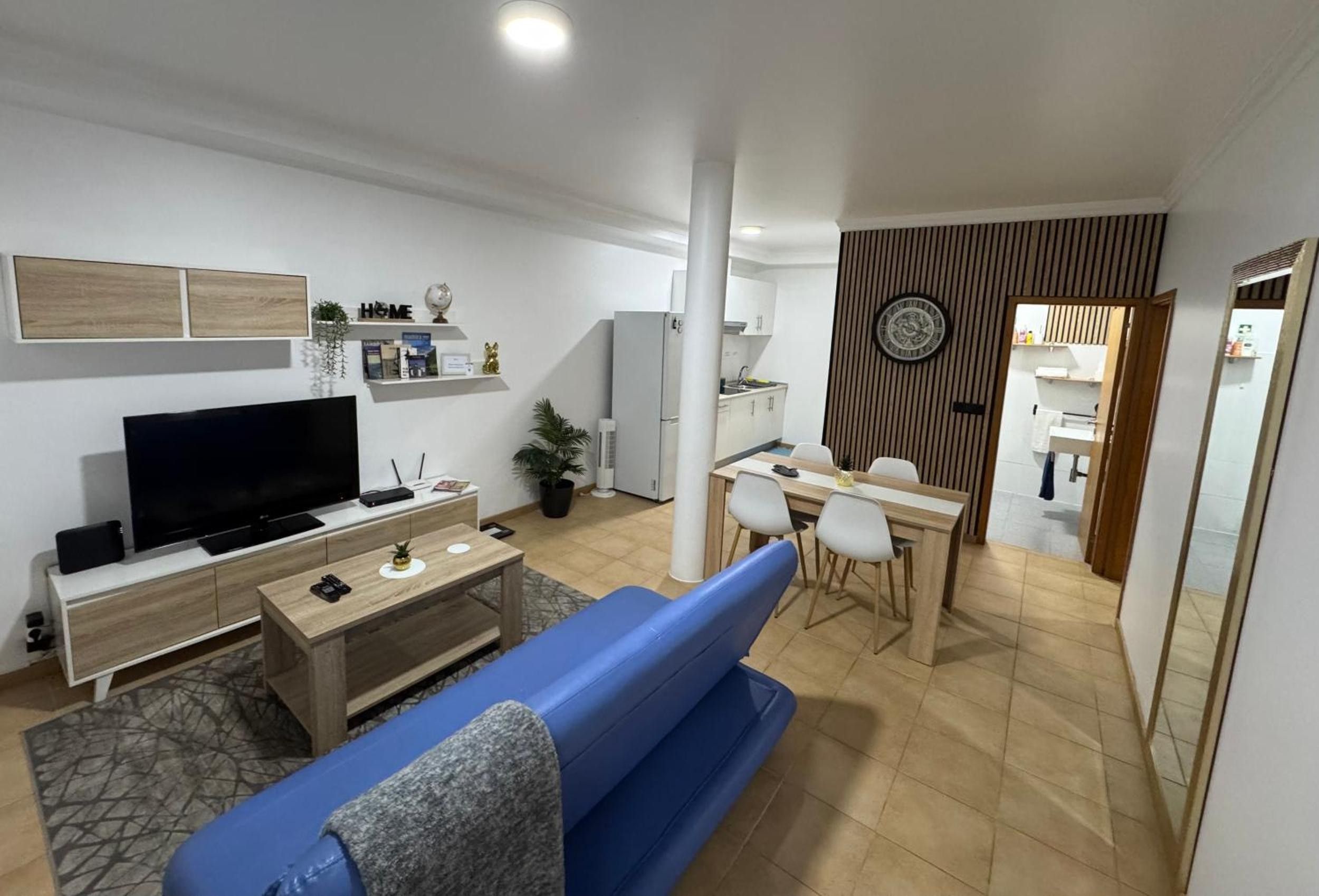 Een moderne woonkamer met een flatscreen televisie, een blauwe bank, een salontafel en een eethoek met een houten tafel en stoelen.