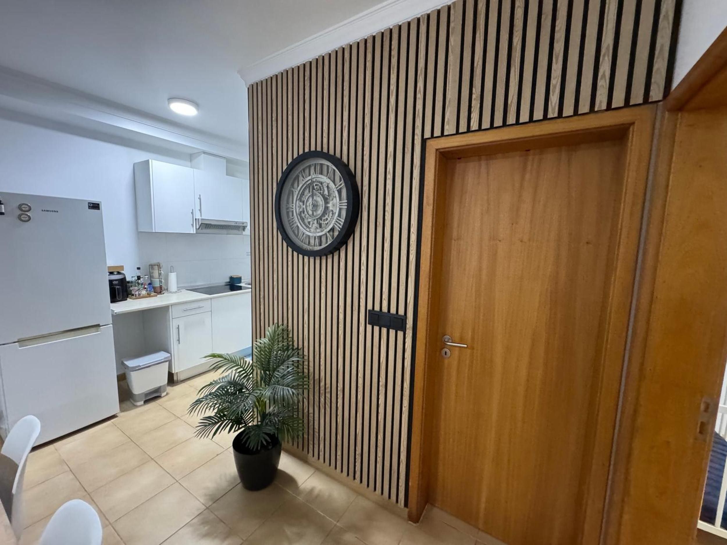 Een moderne keuken met witte kasten, een Samsung koelkast, een houten wandpaneel met een klok en een potplant.