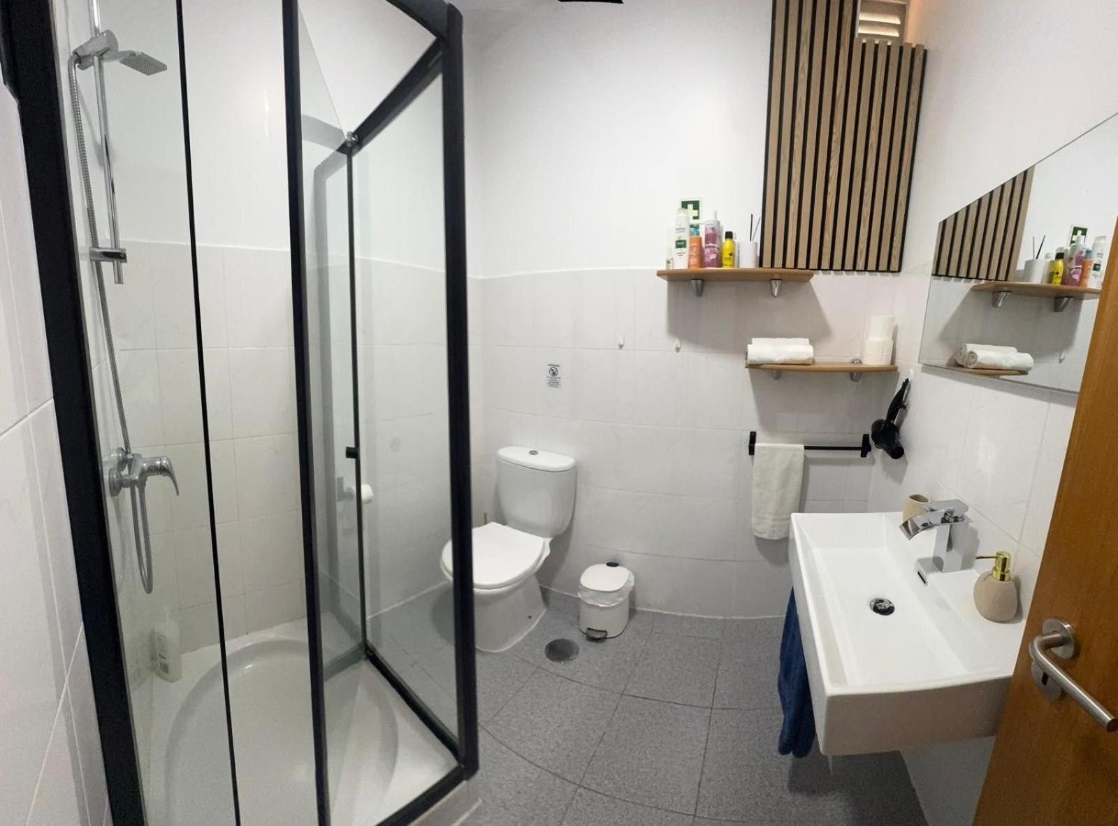 Un baño moderno con ducha envuelta en vidrio, inodoro blanco, lavabo con toalla y artículos de aseo en estantes.
