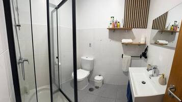 Un baño moderno con ducha envuelta en vidrio, inodoro blanco, lavabo con toalla y artículos de aseo en estantes.