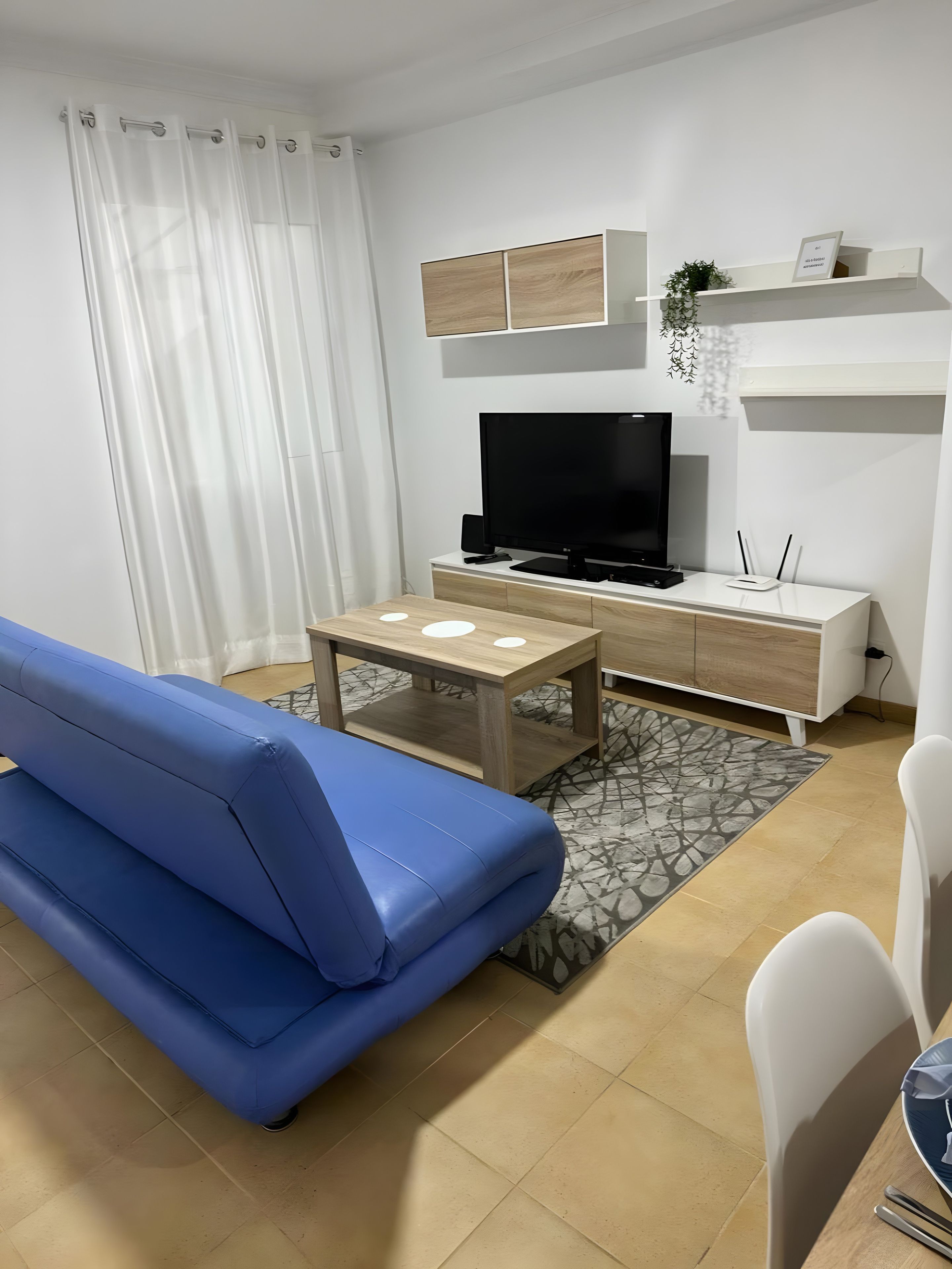 Un salon moderne avec un canapé bleu, une table basse en bois, un téléviseur à écran plat et un coin repas blanc comprenant une table et des chaises.