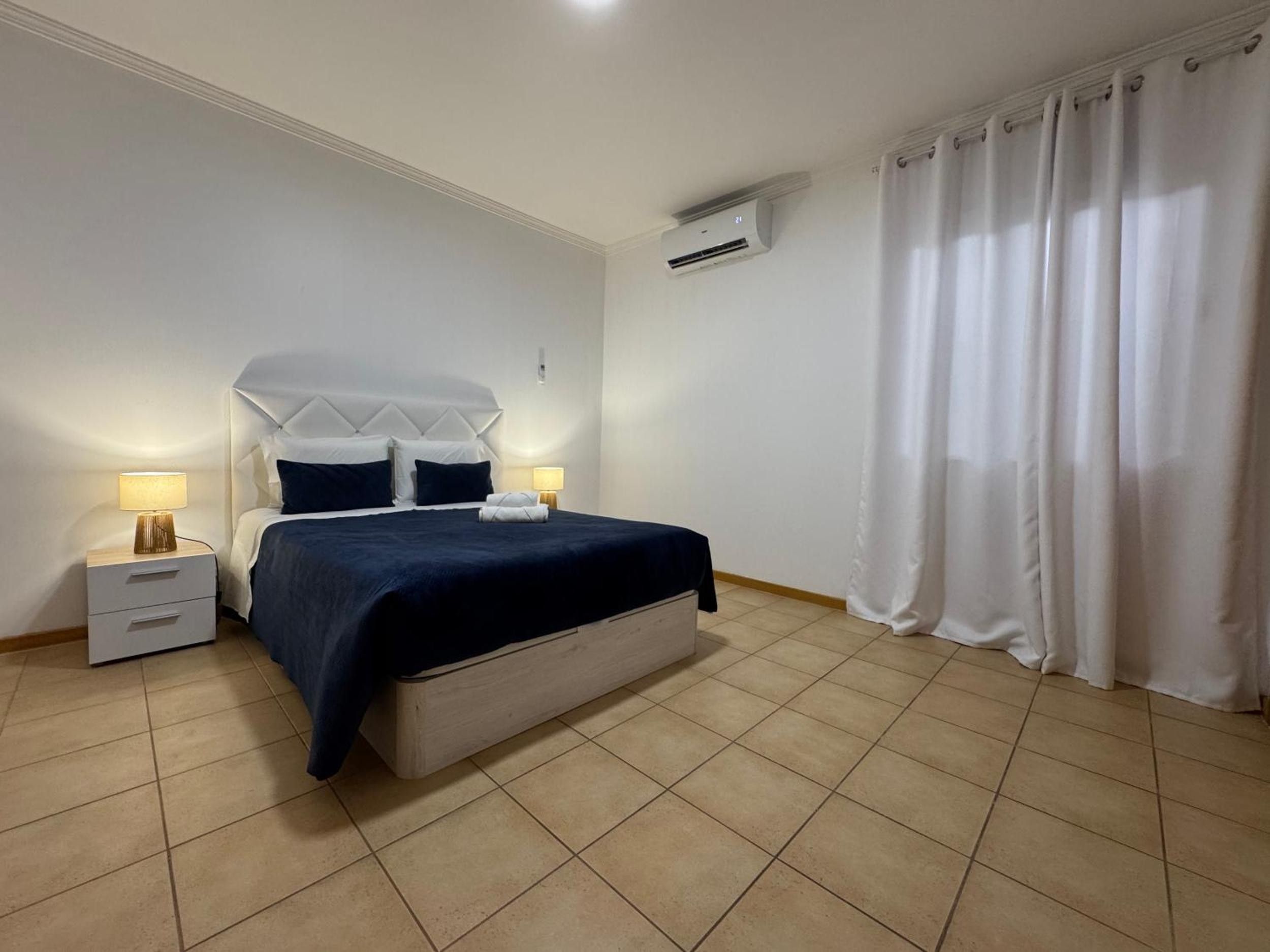 A hotel room with a bed, bedside tables, a lamp, an air conditioning unit, and a curtain.