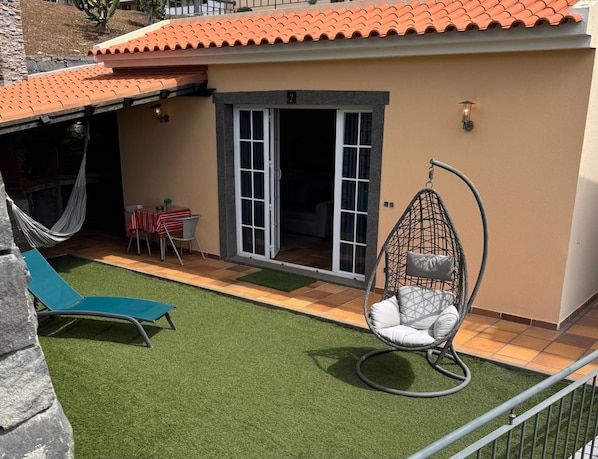 Un patio con césped artificial, una silla colgante, una mesa con mantel a rayas y una hamaca.