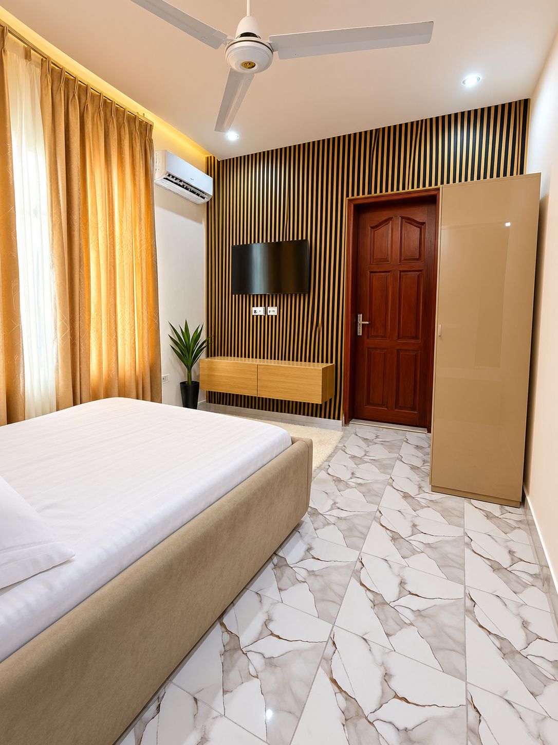 Habitación de hotel con cama, televisor montado en una pared de paneles de madera, armario y ventilador de techo.