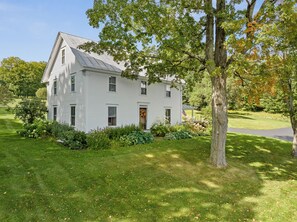 Une maison blanche à deux étages, dotée d’un porche à l’avant, entourée d’une pelouse bien entretenue et d’arbres.
