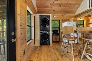 Una cocina compacta con dos lavadoras apiladas, un refrigerador y una mesa de comedor de madera con sillas.