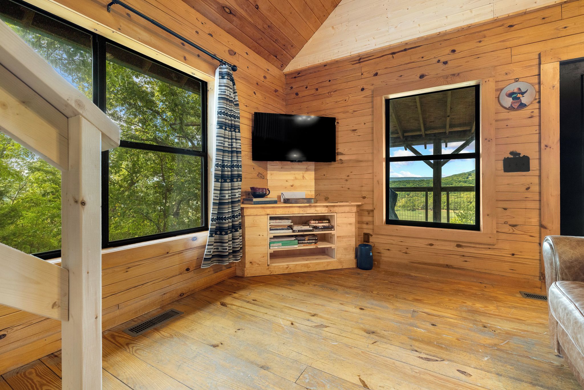 A cabin interior with wooden walls, a television mounted above a wooden shelf, and a large window offering a view of the outdoors.