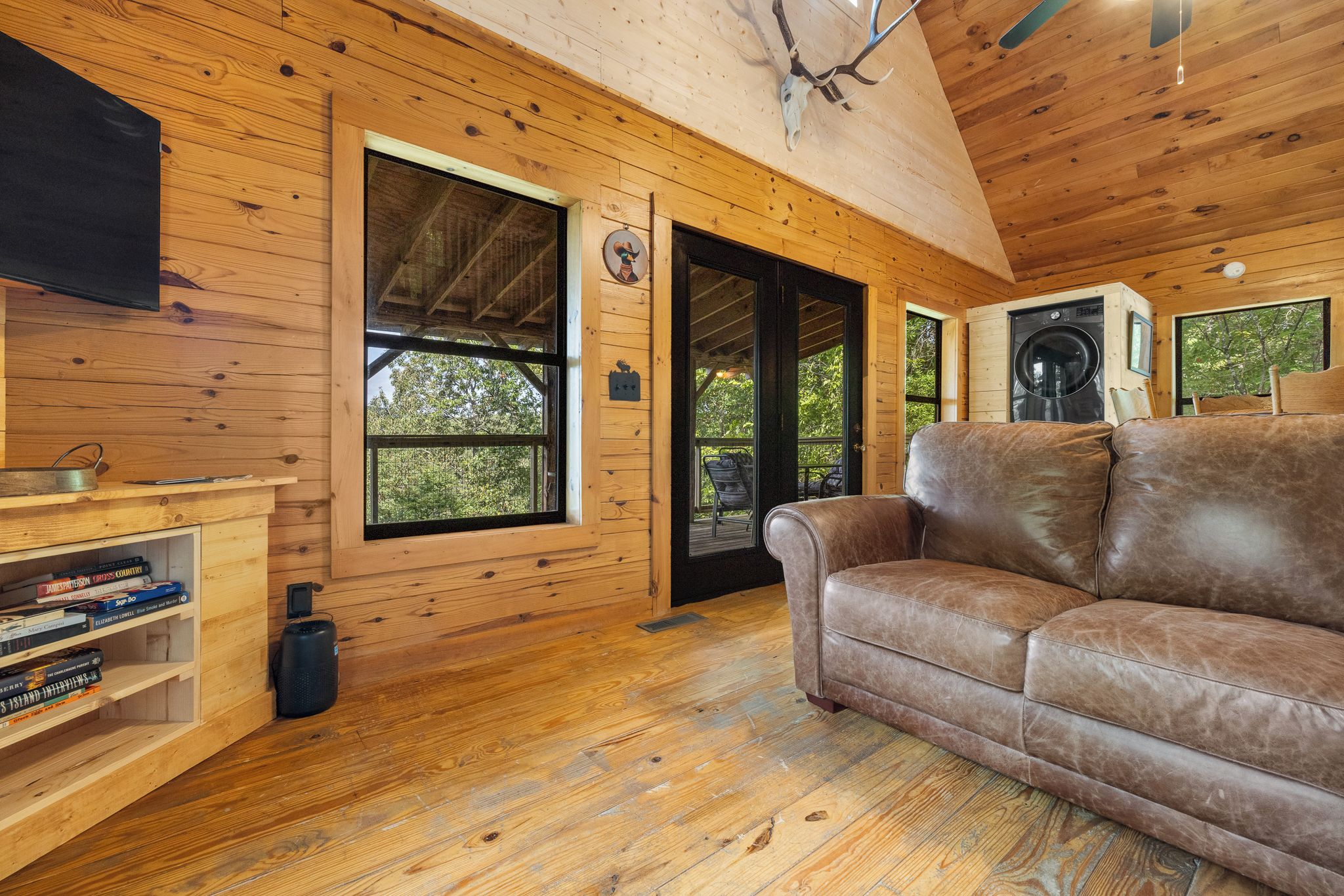 A wooden cabin interior with a leather sofa, a bookshelf, and a large window.
