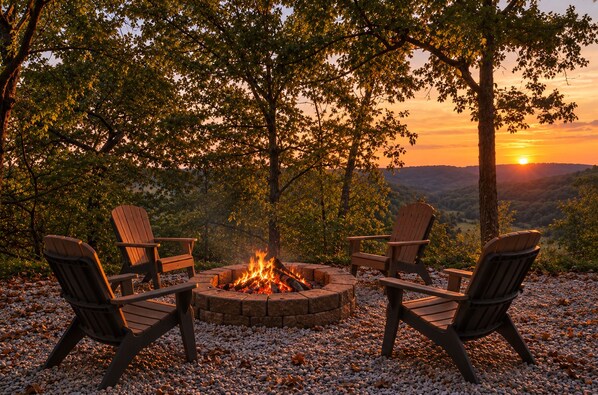 Un hogar con cuatro sillas Adirondack dispuestas alrededor, sobre una base de piedras con árboles de fondo y vista al atardecer.